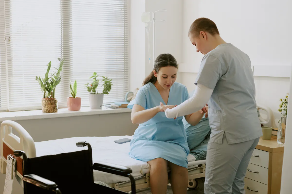 A care worker supporting a young woman with her arm in a sling — a moment of practical, dignified care in a bright, plant-filled room.