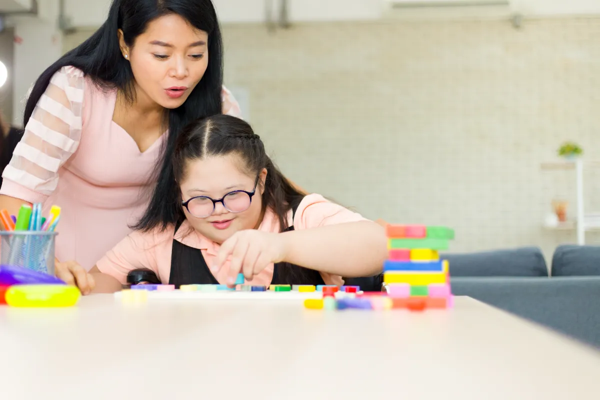 A carer and a young girl building together with colourful blocks — a moment of patient, joyful support.