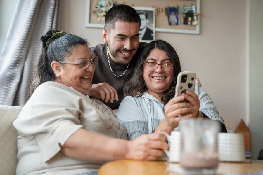 A multi-generational family — grandmother, mother and son — sharing a phone screen together at home, all smiling.